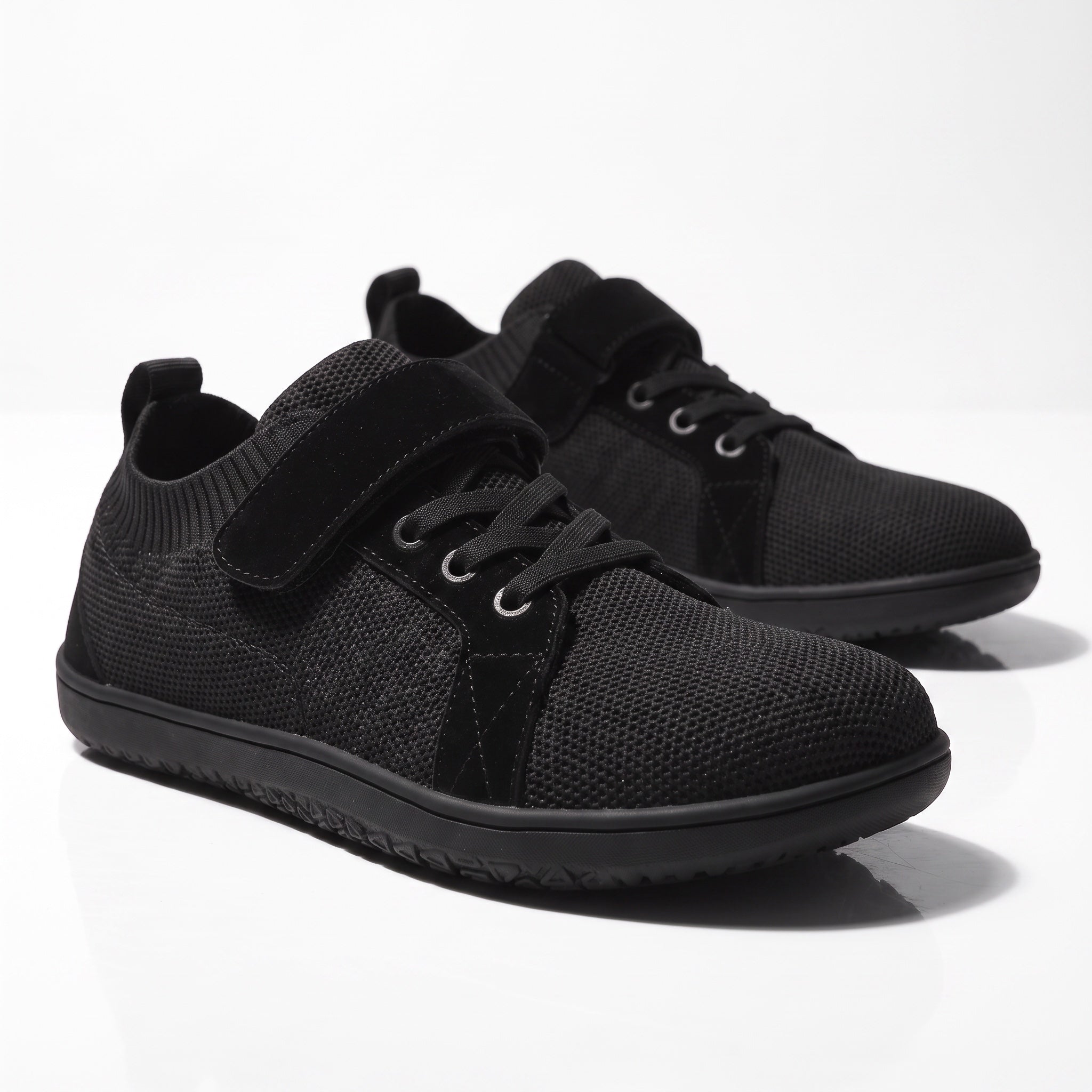 A pair of black low top Children's barefoot shoes with a knit mesh upper, black laces, and hook and-loop strap closures.