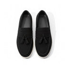 A pair of black slip on loafers with tassel details and white soles.